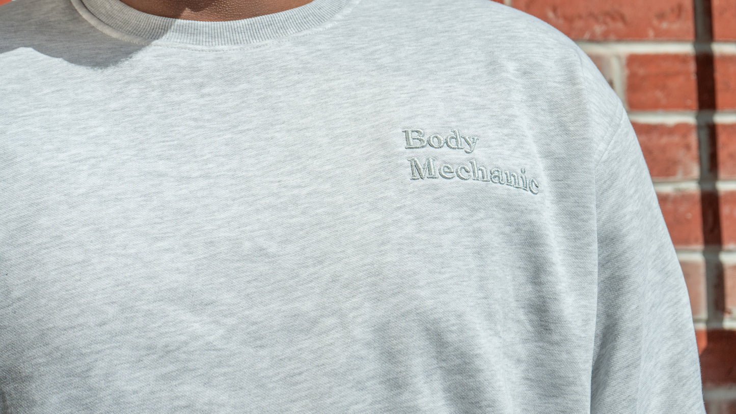Gray sweatshirt with 'Body Mechanic' text worn by a person, against a brick wall background.