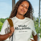 Person wearing a white t-shirt with 'Body Mechanic' text, standing outdoors.