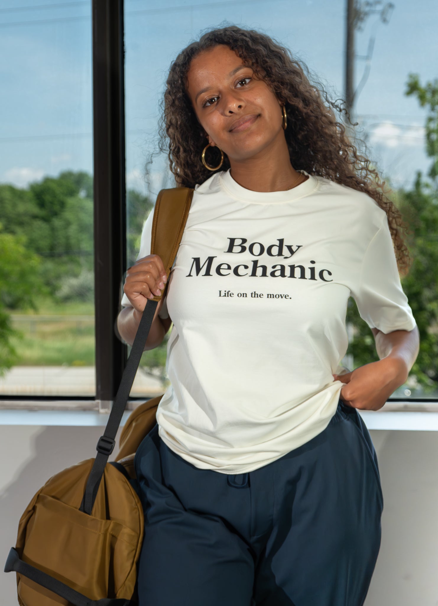 Person wearing a 'Body Mechanic' t-shirt with a backpack, standing by a window with a view of trees.