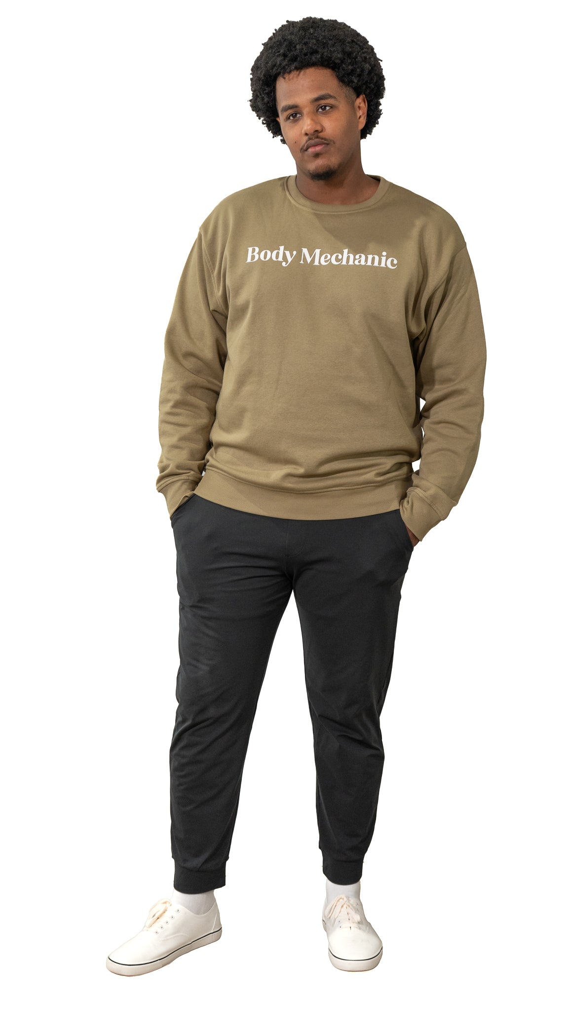 Man wearing a beige sweatshirt with 'Body Mechanic' text, black pants, and white shoes on a white background