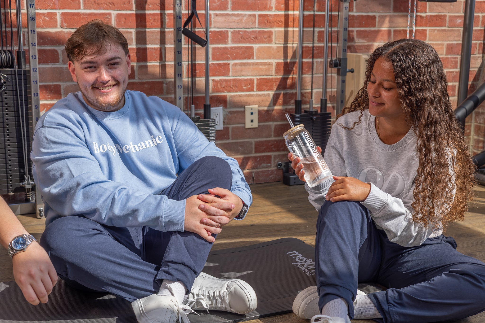Two people sitting on a mat in a gym, one holding a water bottle.
